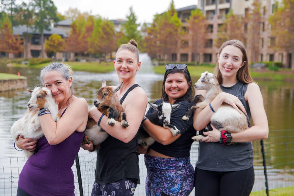 A group of women smiles at the camera from the front lawn of The Woodlands Resort. Each is dressed in yoga or athletic clothes, and each is holding a young goat.