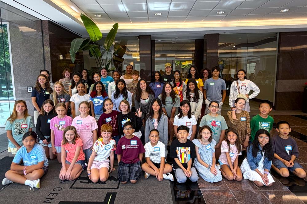 A diverse group of about 45 students (girls and boys, ages 9-18) pose for a group photo inside the front entrance of an office building. Everyone is dressed casually and wearing a sticker name tag for the Young Makers Market workshop.