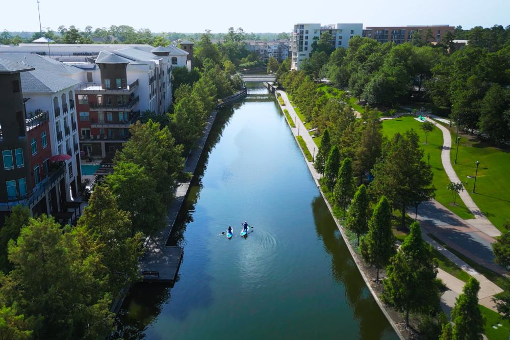 The Woodlands Waterway stretches past apartments at Riva Row and lush green pathways. A couple of kayakers float on the middle of the channel.