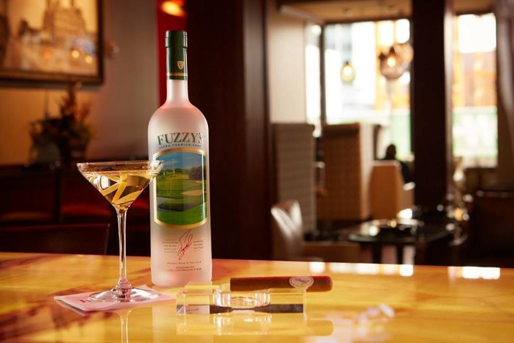 A frosted bottle of Fuzzy's vodka sits atop a light wood counter at BLEND Bar. On its left is a full martini glass with a twist of lemon peel. On its right, a cigar. The lounge area is out of focus behind the counter.