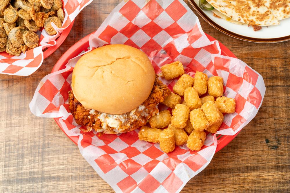 A fried chicken sandwich (buffalo flavored with a creamy white sauce) served with tater tots in a red-and-white checkered basket.