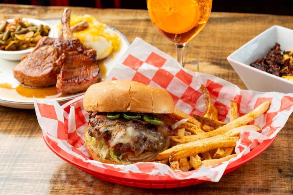A spread of Lankford's dishes: a burger topped with bacon and roasted jalapenos; a glazed pork chop, and a cup of loaded mashed potatoes. With the meal is an alcoholic drink with an orange wedge.
