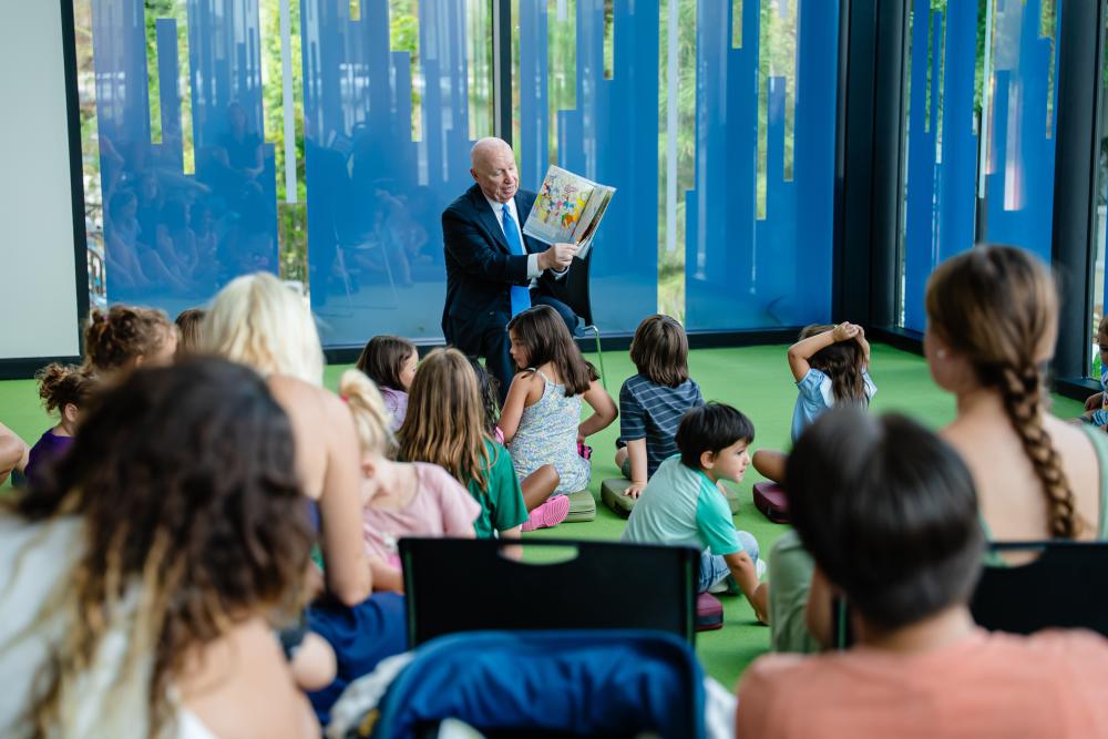 During the grand opening of the library, Kevin Brady reads the children's book, "House Mouse, Senate Mouse." About a dozen children sit on the floor in front of him (their backs to the camera). The camera peeks through the heads of parents sitting behind them.