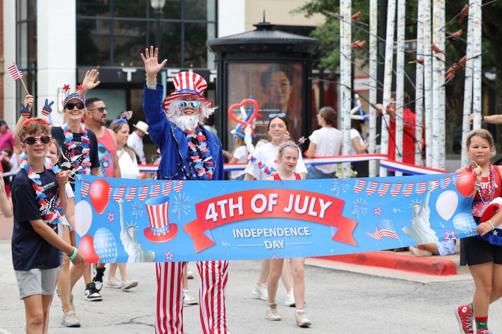 The Fourth of July Parade at Market Street features a performer dressed as "Uncle Sam" followed by a bunch of local families, all dressed in red, white, and blue, some waving banners or American flags. The two children in front carry a sign that reads, "4TH OF JULY | INDEPENDENCE DAY," surrounded by the Statue of Liberty, and RW&B balloons.