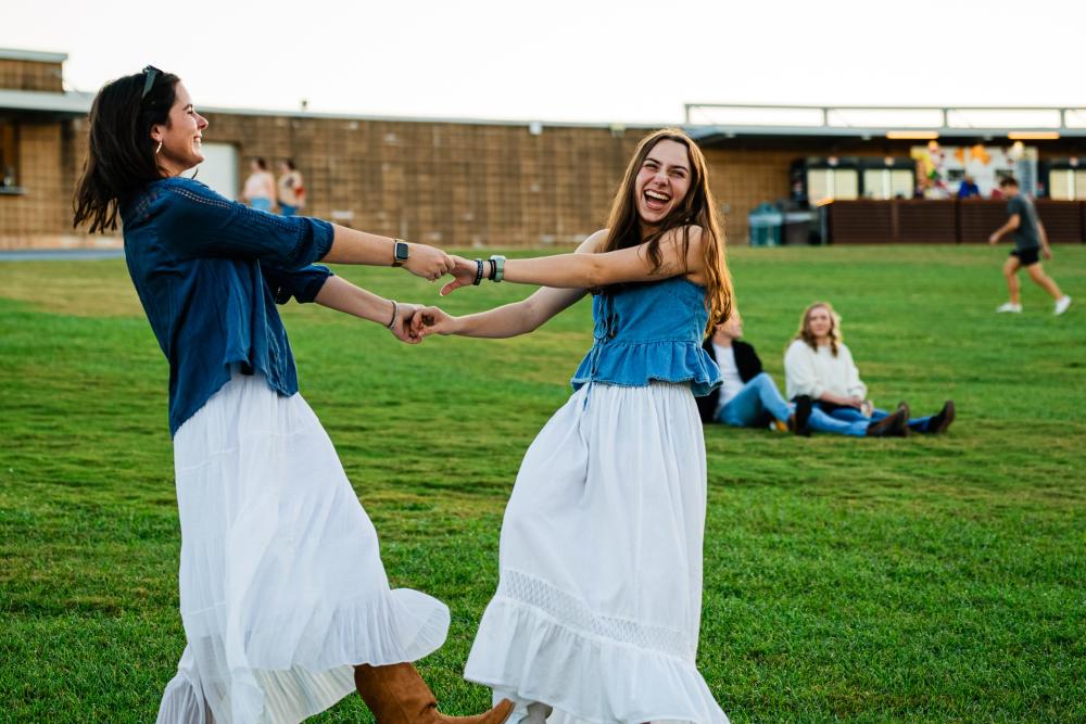 Two young women hold hands and spin on the lawn at The Pavilion. Both are wearing denim tops or jackets with long, casual white skirts and cowgirl boots.