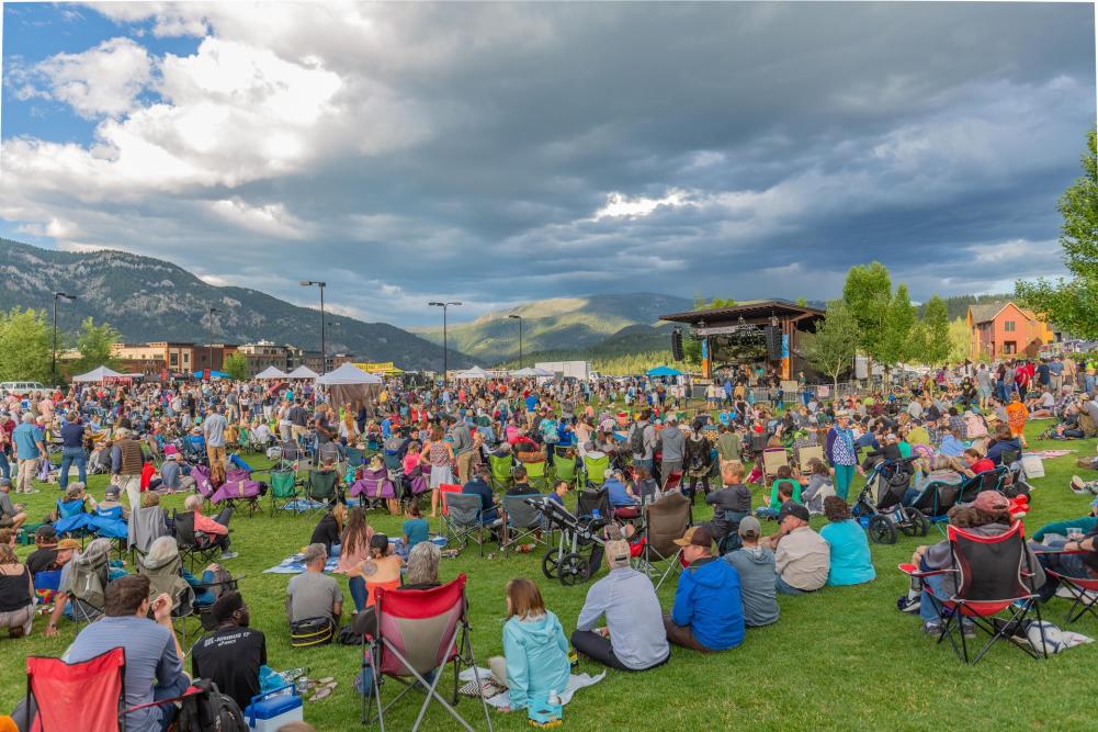 A large crowd gathers on a grassy field for an outdoor event, with a stage in the background and mountains under a cloudy sky.