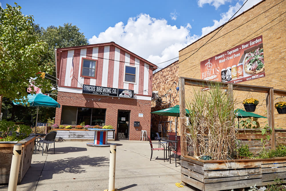A view of the red-and-white striped Bircus Brewing Co buiding with a front patio featuring tables, green umbrellas and hanging plants