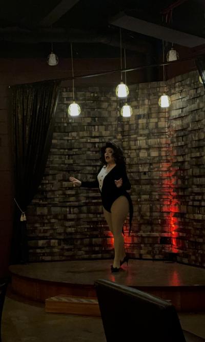 A drag performer stands on a small stage during Huntsville's Legendary Drag Brunch, entertaining guests beneath warm pendant lights in an intimate venue with a rustic wood backdrop and dramatic red accent lighting.