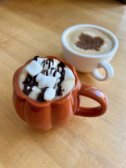 Two lattes, one in an orange mug with marshmallows and chocolate on top, and one in a white mug with a maple leaf design on top, sit on a wooden table.