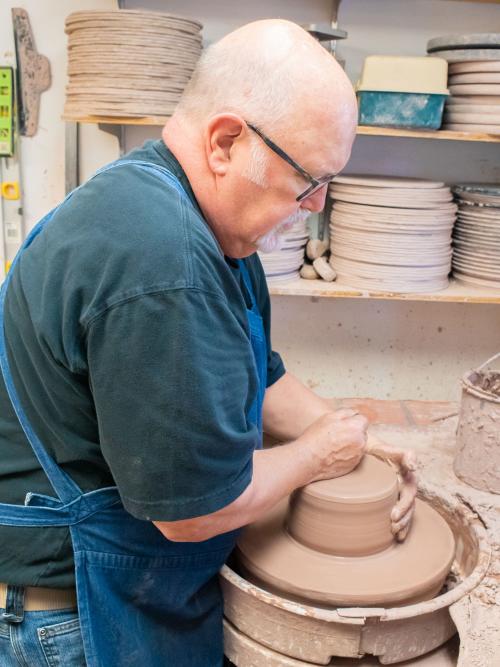 A man stands over a pottery wheel as a large piece of clay is being centered and shaped with his hands.