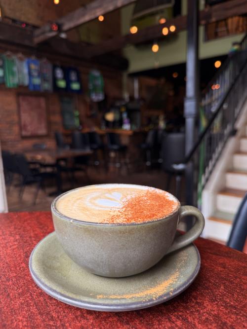 A latte topped with foam art and a dusting of cinnamon sits in a ceramic cup and saucer at Bus Stop Coffee. The drink rests on a red tabletop, with the café's warm interior featuring exposed brick, wooden beams, string lights, and a staircase softly blurred in the background.