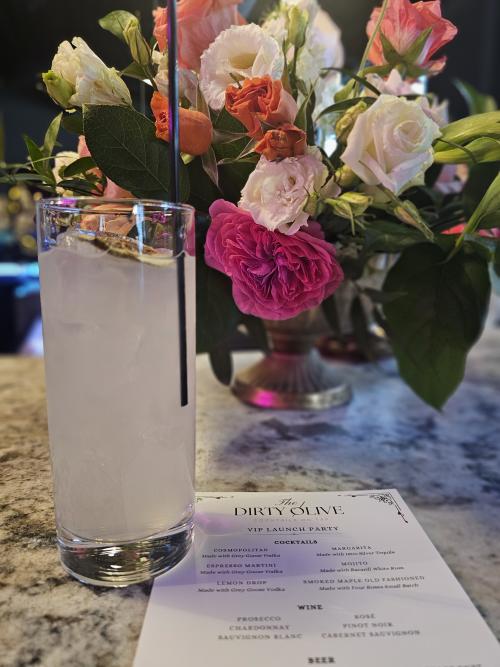 A cocktail sits on a marble bar beside a floral arrangement and a VIP launch party menu at The Dirty Olive in Huntsville, Alabama. Soft lighting, colorful flowers, and an upscale cocktail lounge atmosphere create an elegant and inviting scene.