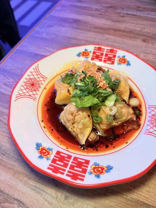 A plate of chili oil dumplings from Dragon Alley in Huntsville, Alabama, topped with fresh cilantro, green onions, and crispy fried shallots, served in a vibrant red chili oil on a decorative dish.
