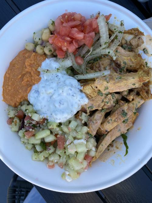 An overhead view of a Mediterranean bowl from Fresko Grille in Huntsville, Alabama, featuring grilled chicken, cucumber quinoa salad, hummus, tzatziki, marinated chickpeas, diced tomatoes, and seasoned onions in a white bowl.