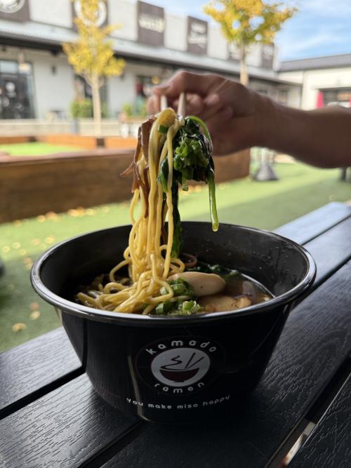A bowl of ramen from Kamado Ramen at Stovehouse in Huntsville, Alabama, with chopsticks lifting noodles above a rich broth filled with greens, mushrooms, scallions, and a soft-boiled egg. The bowl sits on an outdoor patio table with the Stovehouse courtyard blurred in the background.