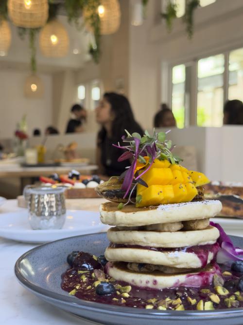 A colorful stack of pancakes topped with fresh mango, bananas, blueberries, berry compote, pistachios, and microgreens is served at Loonas Café in Huntsville, Alabama. Warm lighting, hanging greenery, and guests dining in the background create a bright and cozy brunch atmosphere.