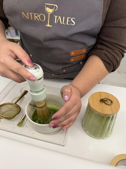 A Nitro Tales employee prepares matcha by whisking bright green tea in a ceramic bowl on a white countertop, with traditional matcha tools and powder nearby. The close-up captures the hands-on process of making matcha inside the café.
