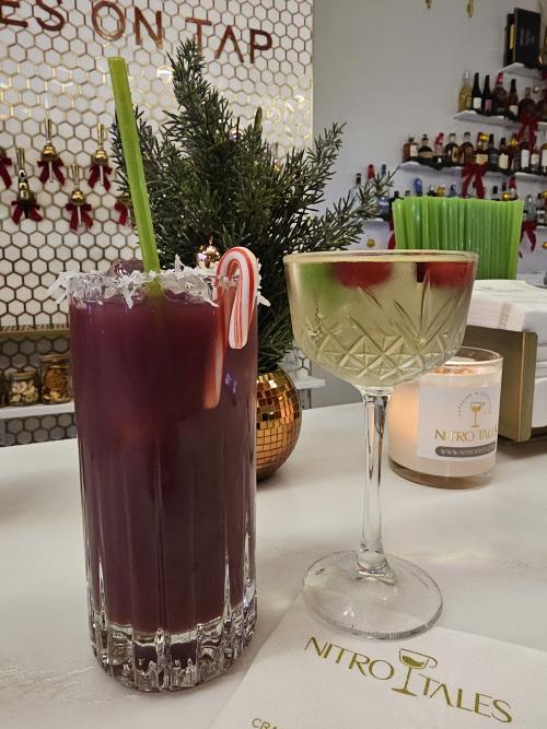 Two seasonal drinks sit on the counter inside Nitro Tales in Huntsville, Alabama, including a deep purple cocktail garnished with a candy cane and coconut rim beside a festive martini-style drink. Holiday décor, candles, and shelves of bottles in the background create a cozy and upscale atmosphere.
