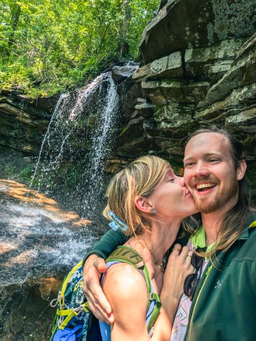 A couple takes a selfie in front of a small waterfall, with one person kissing the other on the cheek. Surrounded by lush greenery and rock formations, the scene captures a joyful moment during an outdoor hike on Monte Sano Mountain.