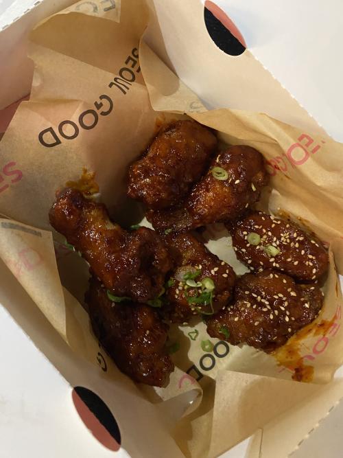 An order of Korean fried chicken wings from Seoul Good in Huntsville, Alabama, coated in a glossy sauce and topped with sesame seeds and sliced green onions, served in a paper-lined takeout basket.