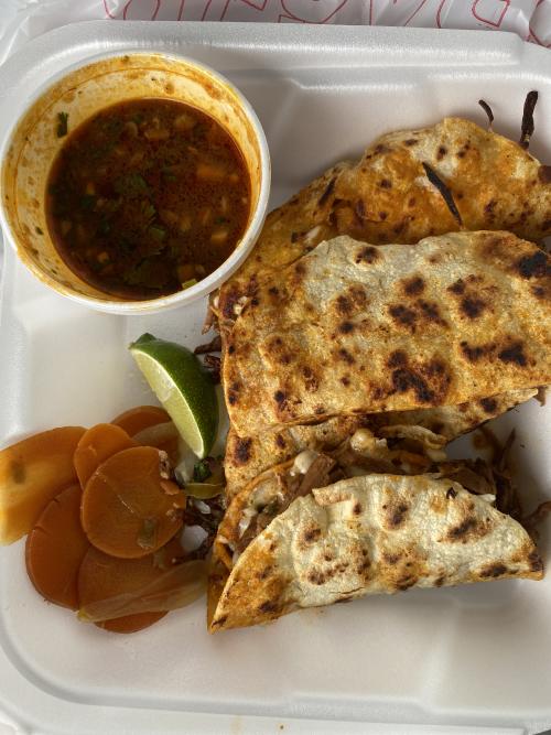 A takeout container from Taqueria El Cazador featuring several crispy birria tacos served with a cup of rich consommé, pickled carrots and onions, and a fresh lime wedge. The tacos are filled with shredded meat and melted cheese and photographed from above.