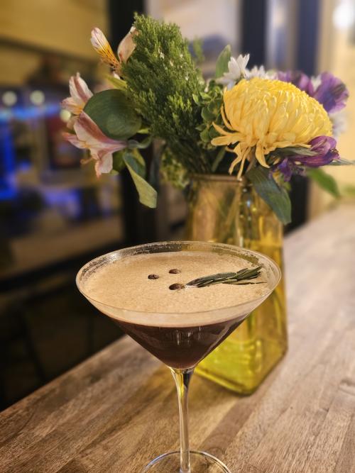 An espresso martini topped with creamy foam and coffee beans sits on a wooden bar beside a colorful floral arrangement at The Camp in Huntsville, Alabama. Warm lighting and a softly blurred background create a cozy and relaxed cocktail lounge atmosphere.