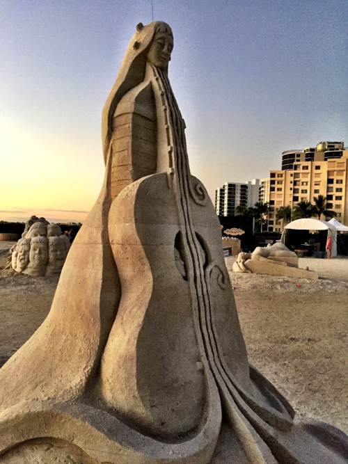 Sand sculpture showing a guitar standing straight up. At the top of the guitar, instead of what's usually there, there's a woman's head. Her body flows down behind the guitar