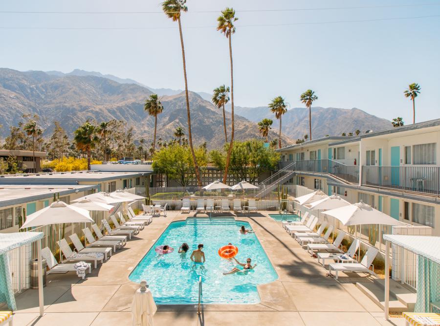 An outdoor swimming pool at a hotel with people relaxing in and around the pool, surrounded by palm trees with mountainous scenery in the background. Sun loungers and umbrellas line the poolside, and a two-story hotel building flanks the pool area.
