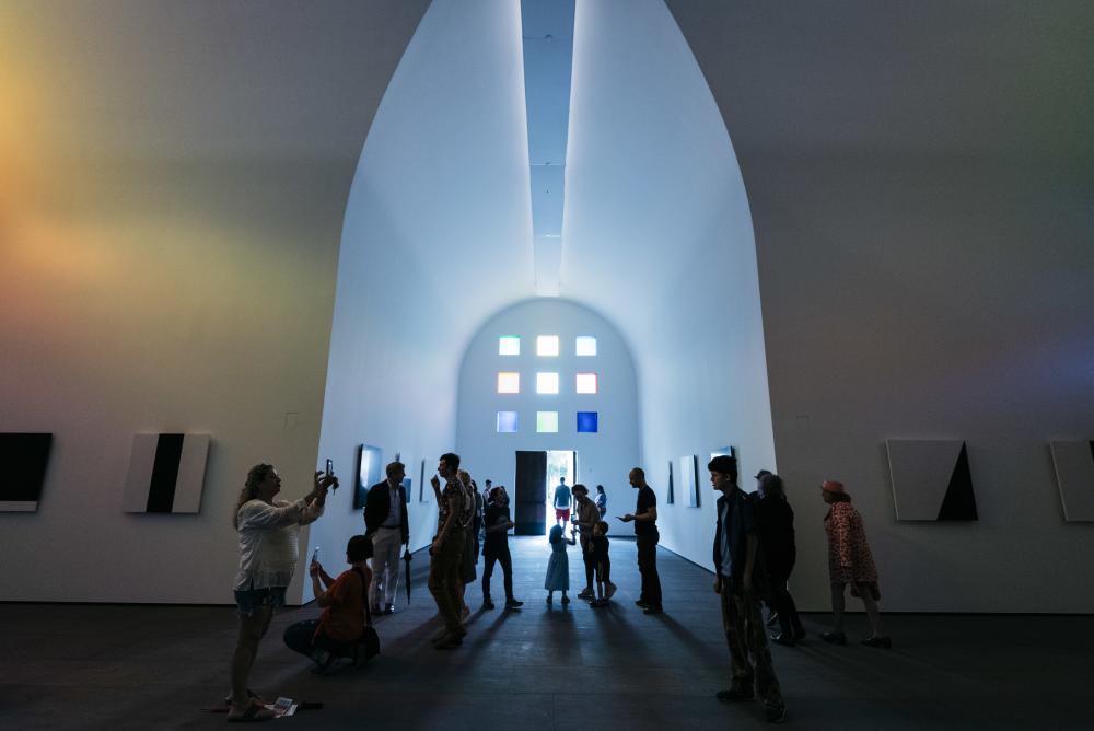 The inside of the Ellsworth Kelly exhibit at the Blanton Museum with visitors walking around and a large light source coming from the stained glass windows on the front of the building.