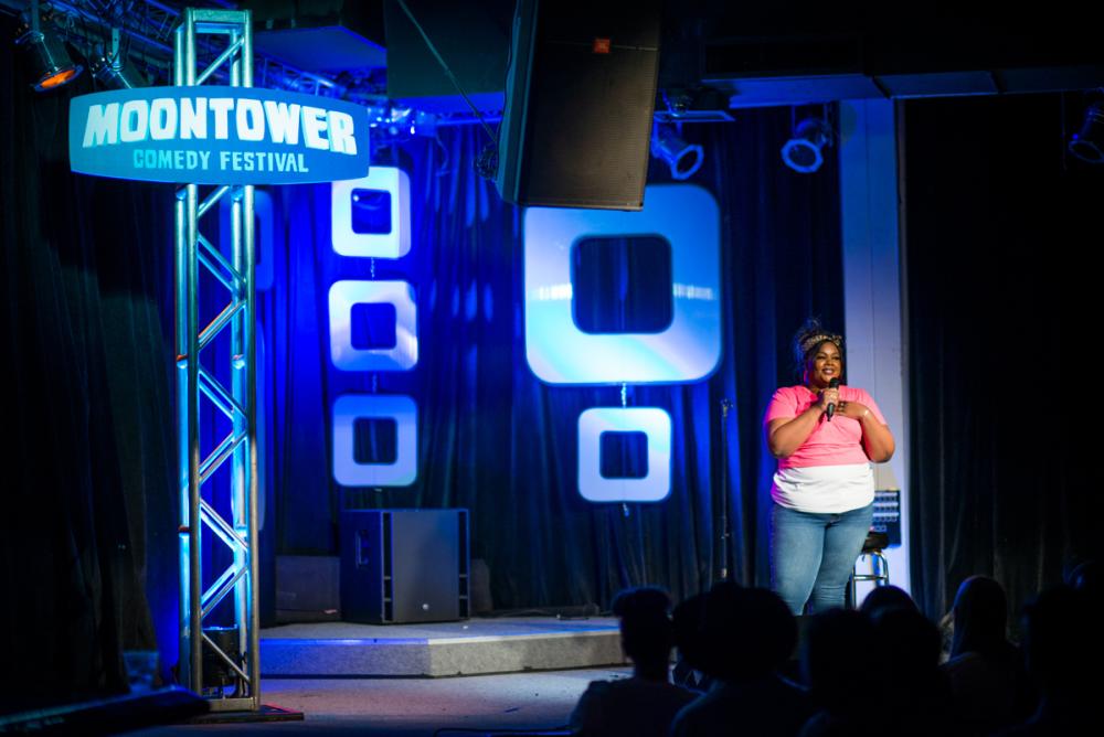 Nicole Byer performs on stage next to sign reading Moontower Comedy Festival