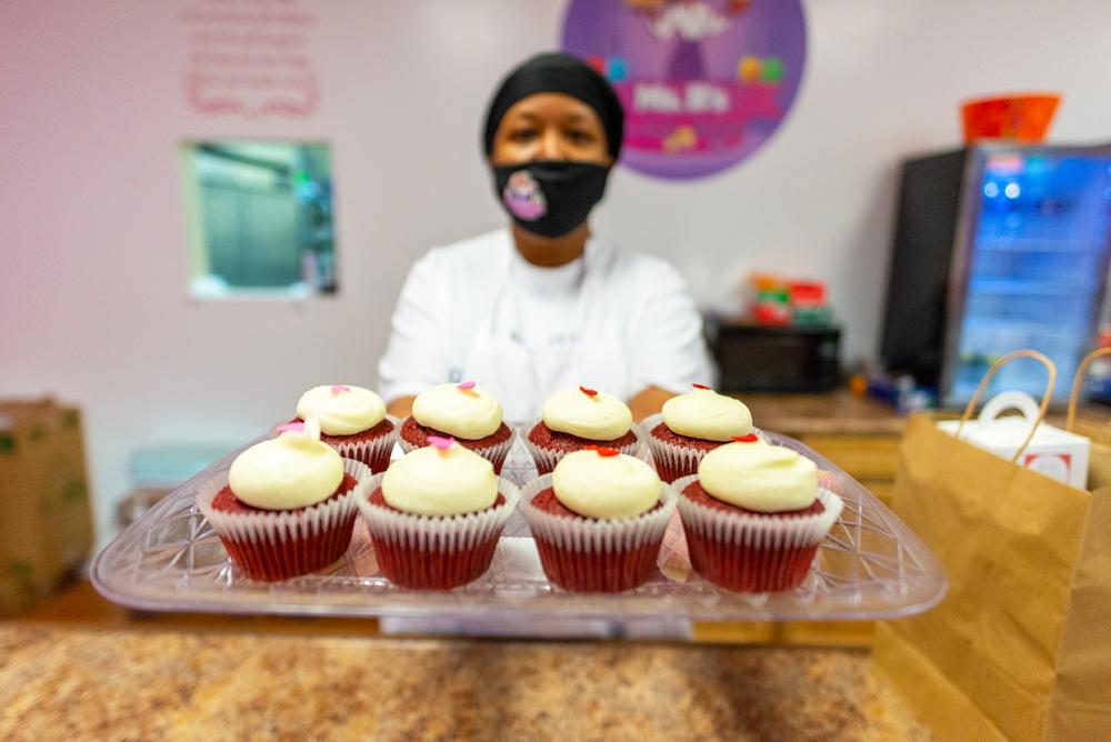 Lady with a mask poses with a tray of red velvet cupcakes