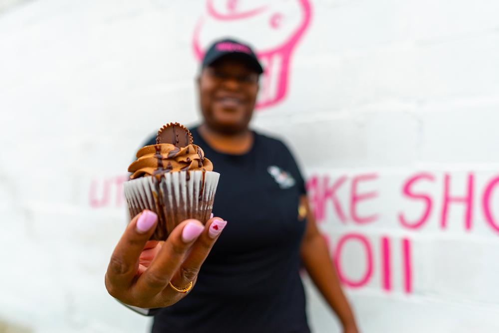 A woman poses in front of her cupcake shop holding out a chocolate cupcake