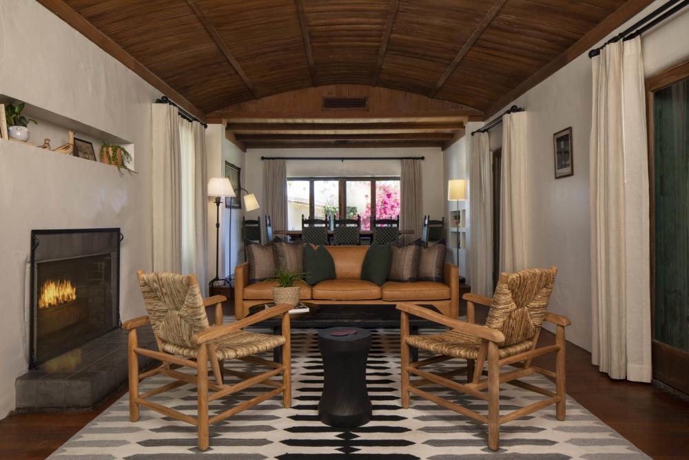 A cozy living room with a fireplace, a leather sofa flanked by wicker armchairs, a black and white patterned rug on the floor, and exposed wooden beams on the ceiling. A window in the background offers a view of pink flowers outside.