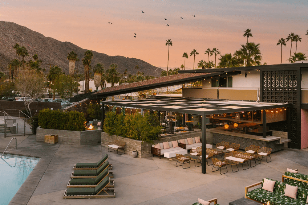 Outdoor patio area of a modern restaurant with lit string lights at dusk, poolside lounge chairs in the foreground, surrounded by mountainous terrain and palm trees under a twilight sky with birds flying.