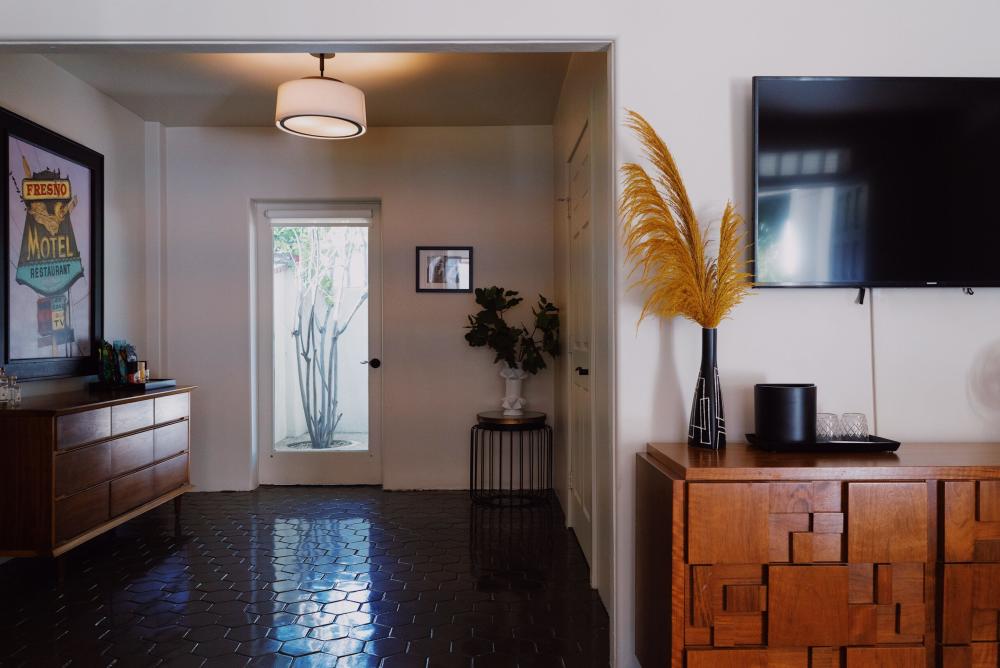 A well-decorated room featuring a dark hexagonal tile floor, wooden mid-century modern-style dresser, black flat-screen TV on the wall, and a round ceiling light fixture. To the right, a tall potted plant with yellow leaves by a black vase sits on a wooden instrument case, and to the left, a doorway leads to a bright area with obscured glass, showcasing a silhouette of a tree outside. Decorative elements include a framed poster, a small photo on the wall, and a circular table with a vase and greenery.