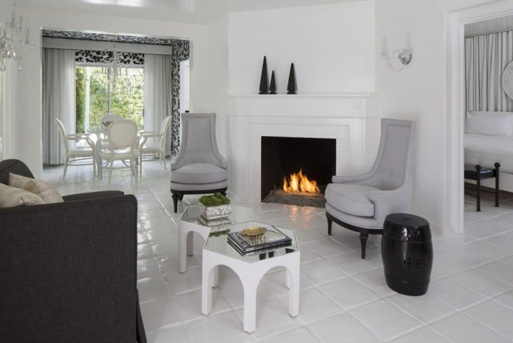 A modern living room with a lit fireplace, flanked by two high-back chairs, a glass-topped coffee table, and a view into a dining area with white chairs and curtains.
