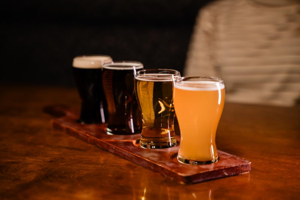 A beer flight is lined up across the frame from darkest to lightest. A young woman sits behind it.