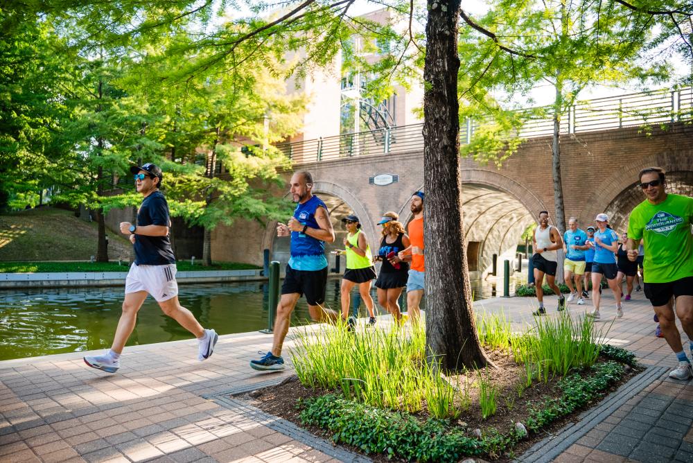 A group of runners jog under the Waterway Avenue Bridge and past the viewer. There are men and women, mostly in the 30s-40s age range. Green grass and trees abound.