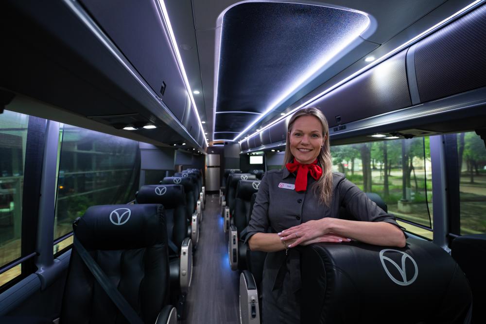 The interior of a Vonlane luxury motor coach features 2 rows of plush, reclining leather seats and low lighting. The attendant on board, a young blonde woman wearing a gray uniform and red bow around her neck, smiles at the viewer.