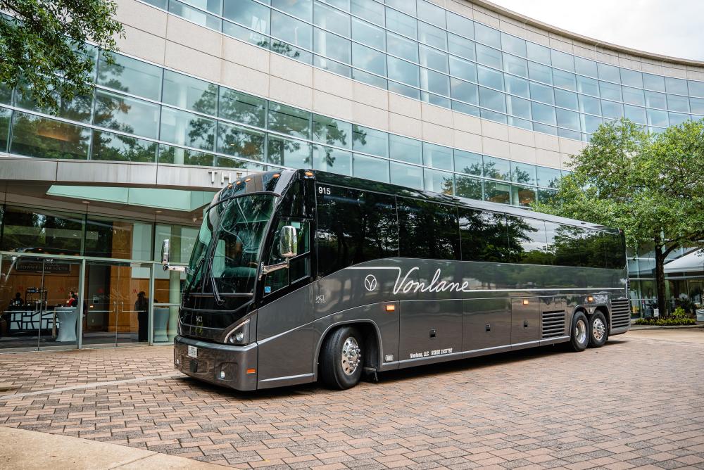 A long silver bus bearing the "Vonlane" name and logo is parked in front of The Woodlands Waterway Marriott Hotel & Convention Center. The front of the hotel is all glass windows.