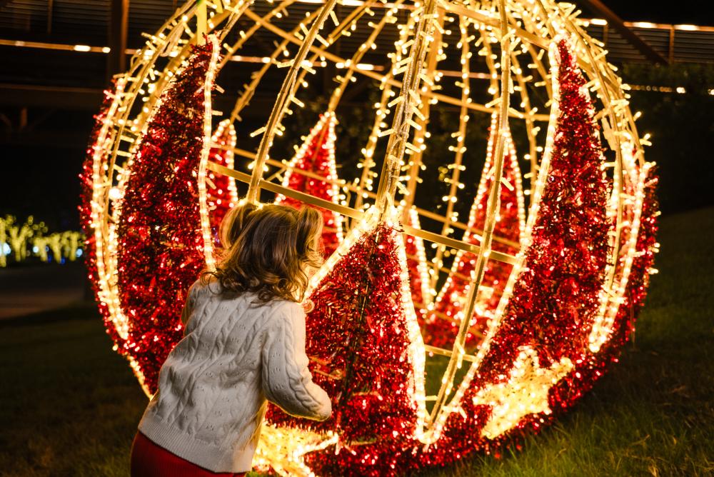 A toddler in a white sweater and pigtails peers through a light display shaped like a giant Christmas ornament during Holiday on The Waterway in The Woodlands, TX.