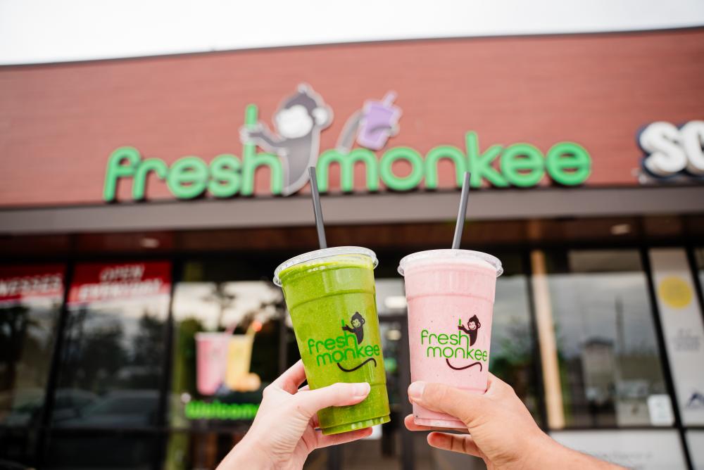 A young couple (off-screen) holds up their shakes from Fresh Monkee in front of the shop. One shake is green, the other pink. The "fresh monkee" font is all lowercase letters, all bright green, with a logo of a monkey holding a purple shake with its tail.