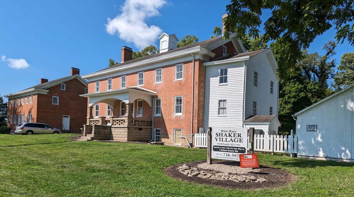 Image is of the White Water Shaker Village welcome sign with the Dwelling House behind it and the Meeting House to the left.
