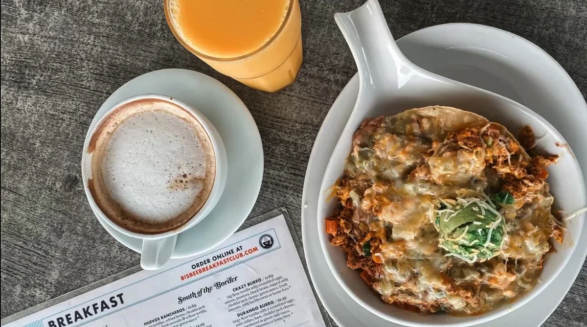 a cup of coffee, a glass of orange juice, and a plate of a cheesy breakfast scramble on a table next to a a menu that reads "Bisbee Breakfast Club"