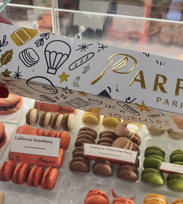 Image of a white box with gold font that reads "Parfait Paris." In the background a variety of macarons can be seen; each a different color. There are red, brown, white, yellow, and green macarons.