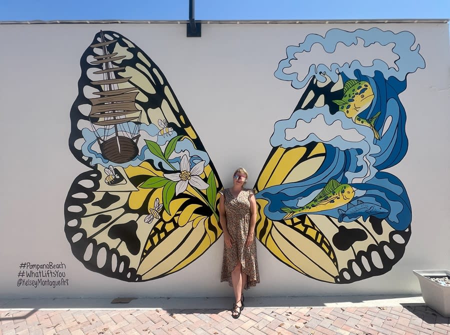 A woman in a flowy light brown dress is posing in front of a mural of butterfly wings.