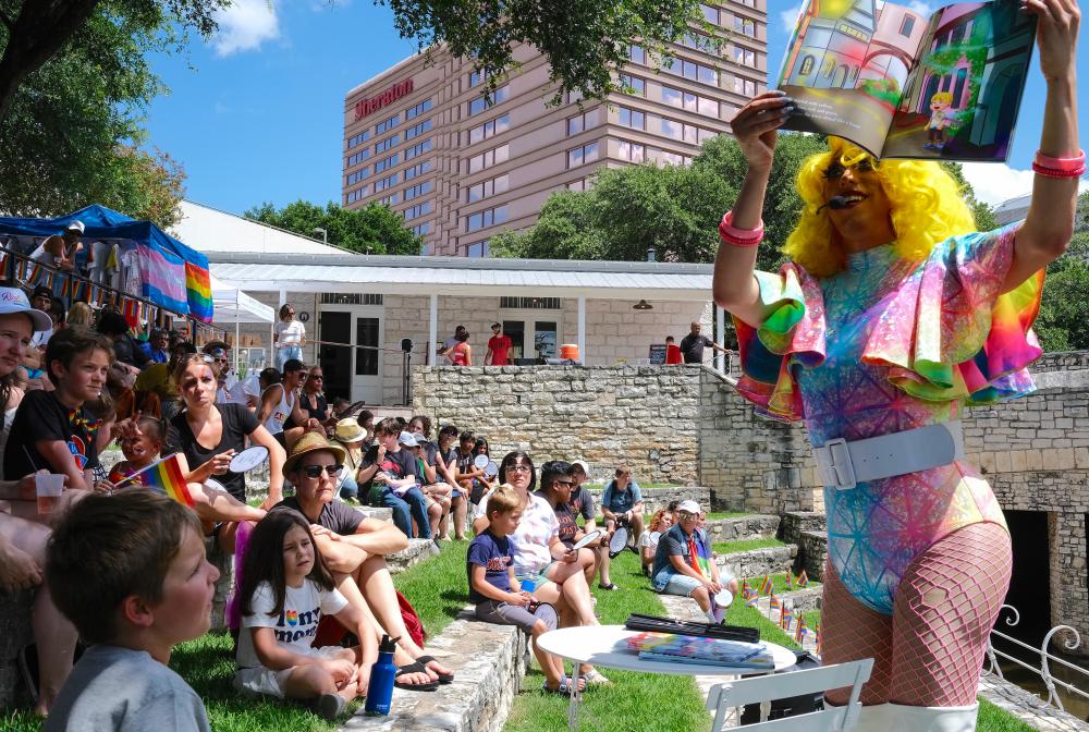 Drag queen decked out in rainbow apparel walking through crowd of children and adults, showing off the illustrations of a children's book.