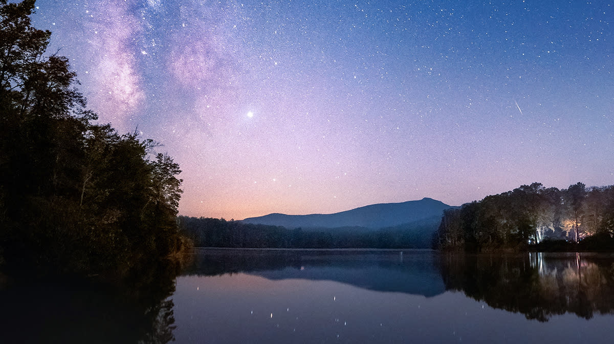 The Milky Way Galaxy can be seen shining above the nighttime silhouette of Grandfather Mountain. The star-filled sky can be seen in the reflection of Julian Price Lake in the foreground.