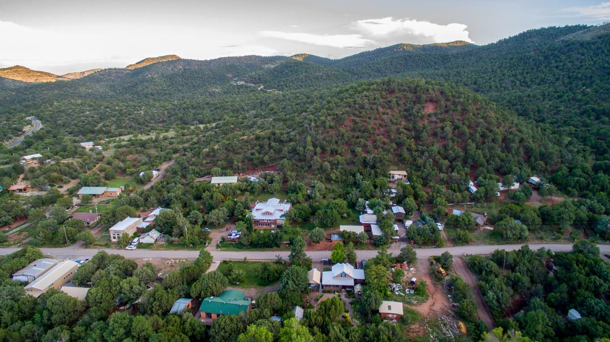 A scenic aerial view of a small town surrounded by lush green hills and trees. Residential houses and buildings are visible amidst the natural landscape, with a road running through the area. The sky is clear with a few clouds.