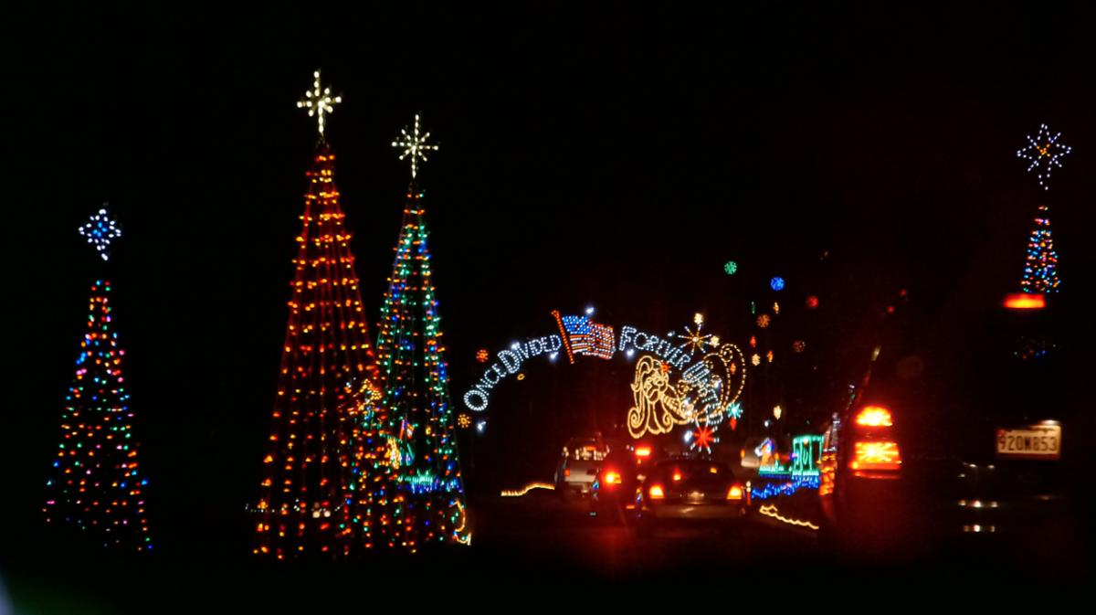A holiday drive through light show is shown at nighttime, with cars lined up next to lit up trees and an archway reads "once divided, forever united".